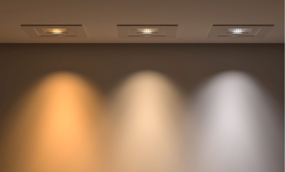 Tre faretti quadrati a incasso nel soffitto che proiettano coni di luce di diversa temperatura cromatica: caldo (giallo-arancio), neutro (bianco tenue) e freddo (bianco brillante), per illuminazione artificiale in una stanza.